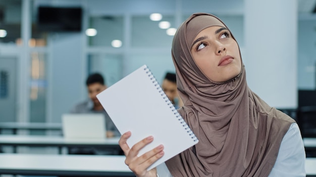 Jeune travailleuse arabe pensive en hijab assise au bureau à l'aide d'un cahier en papier prenant des notes pour créer une nouvelle idée