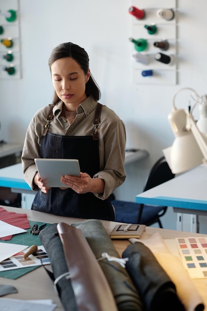 Jeune travailleuse du cuir avec tablette regardant à travers de nouvelles commandes en ligne