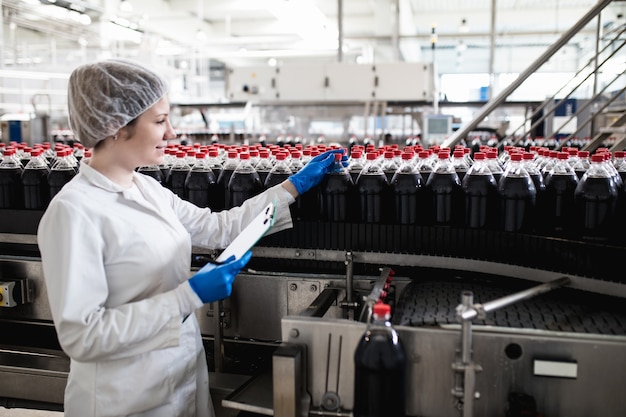 Jeune travailleuse heureuse dans l'usine d'embouteillage vérifiant les bouteilles de jus avant expédition