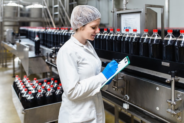 Jeune travailleuse heureuse dans l'usine d'embouteillage vérifiant les bouteilles de jus avant expédition