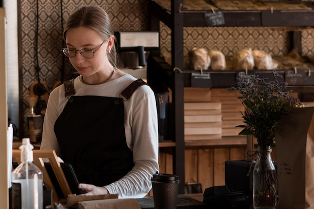 Jeune vendeuse travaillant dans un café ou un café avec un appareil mobile et une caisse enregistreuse