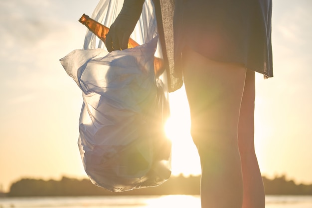 Un jeune volontaire en gants noirs marche avec un sac poubelle le long d'une plage sale de la rivière