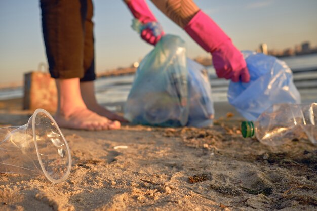 Un jeune volontaire en gants violets marche avec un sac poubelle le long d'une plage sale de la rivière