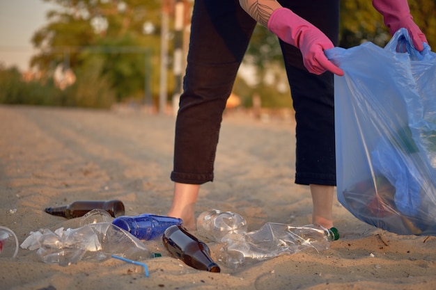 Un jeune volontaire en gants violets marche avec un sac poubelle le long d'une plage sale de la rivière