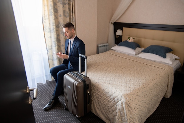 Jeune voyageur d'affaires prospère en costume assis sur le lit et défilement dans le smartphone tout en allant appeler un taxi
