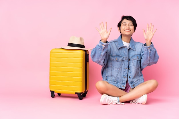 Jeune voyageur femme vietnamienne avec valise assis sur le sol sur le mur rose comptant dix avec les doigts