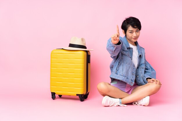 Jeune voyageur femme vietnamienne avec valise assis sur le sol sur le mur rose isolé montrant et levant un doigt