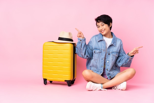 Jeune voyageur femme vietnamienne avec valise assis sur le sol sur le mur rose isolé pointant le doigt vers les latéraux et heureux