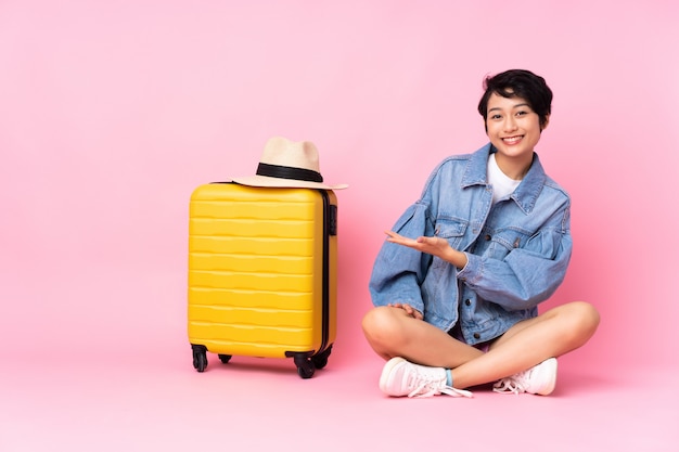 Jeune voyageur femme vietnamienne avec valise assis sur le sol sur mur rose souriant