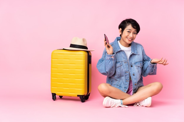 Jeune voyageur femme vietnamienne avec valise assise sur le sol sur le mur rose en gardant une conversation avec le téléphone mobile avec quelqu'un