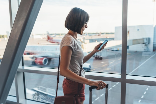 Jeune voyageuse occasionnelle à l'aéroport tenant un téléphone intelligent près des fenêtres de la porte des avions sur la piste