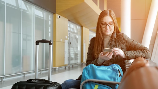Une jeune voyageuse passe l'enregistrement électronique sur son téléphone portable en attendant son vol à l'aéroport. Elle est assise dans le hall avec sa valise et son sac à dos, la lumière du soleil