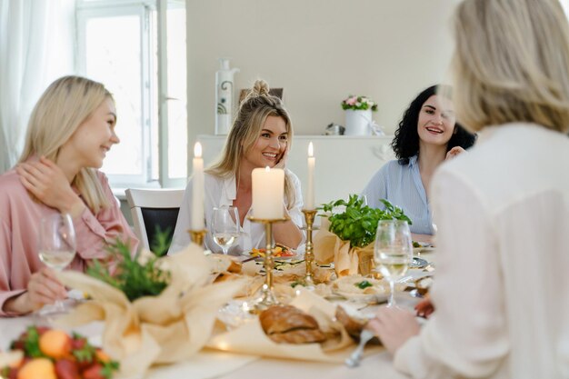 Jeunes amies heureuses riant à la table de fête servies avec de la nourriture et du vin blanc décorés de bougies