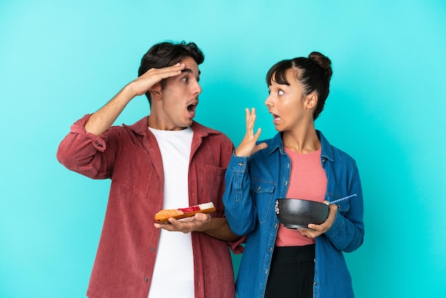 Jeunes amis métis tenant des sashimis et des ramen isolés sur fond bleu avec une expression faciale surprise et choquée