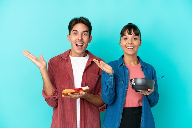 Jeunes amis métis tenant des sashimis et des ramen isolés sur fond bleu avec une expression faciale surprise et choquée