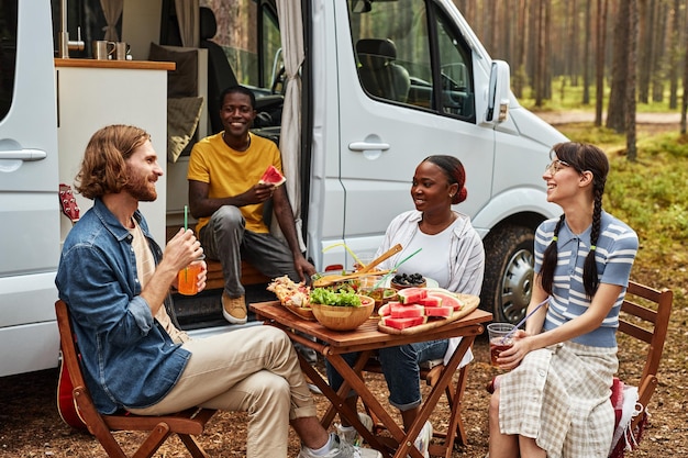 Jeunes amis en train de déjeuner à table lors d'un pique-nique dans la forêt, ils voyagent sur une maison de roues