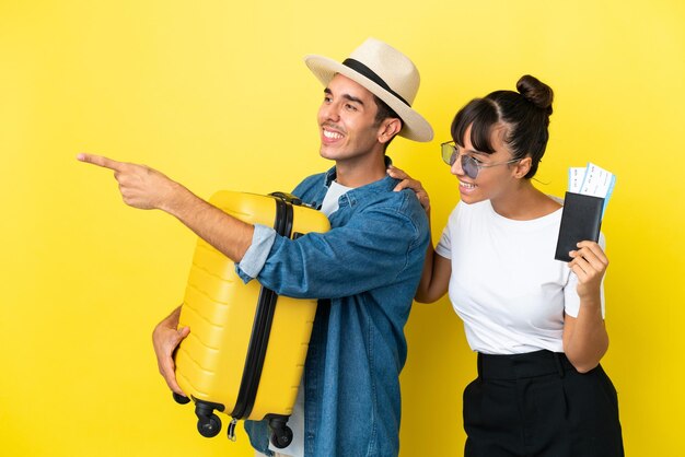 Jeunes amis voyageurs tenant une valise et un passeport isolés sur fond jaune pointant vers le côté pour présenter un produit