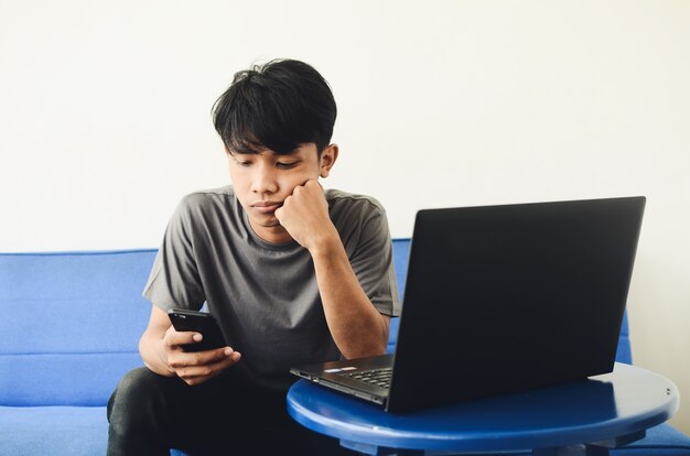 Jeunes asiatiques assis sur le canapé devant l'ordinateur portable à l'aide d'un téléphone portable