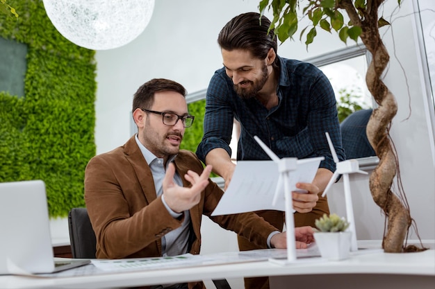 Jeunes collègues d'affaires prospères analysant un projet d'énergie alternative avec des miniatures de turbines éoliennes à la table, et dans une discussion sérieuse, regardant la paperasse.