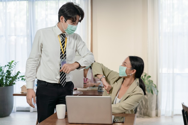 Jeunes collègues asiatiques hommes et femmes portant un masque de salutation avec des coudes qui se cognent pendant l'épidémie de coronavirus COVID-19 au bureau