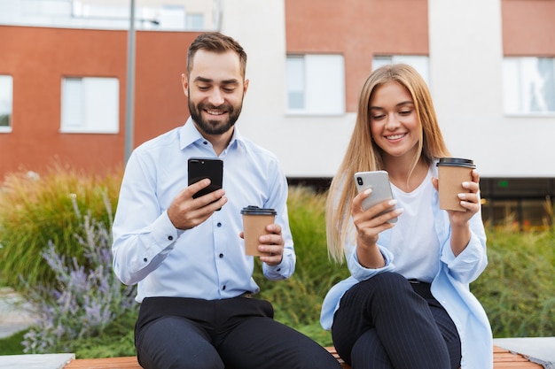 Jeunes Collègues Joyeux Et Souriants, Hommes Et Femmes D'affaires Sont Assis Dehors Dans La Rue à L'aide De Téléphones Portables.