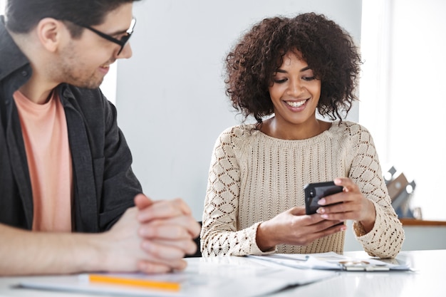 Jeunes collègues joyeux utilisant un smartphone ensemble tout en étant assis près de la table au bureau