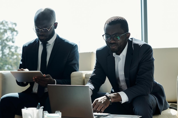 Jeunes collègues masculins afro-américains sérieux utilisant une tablette et un ordinateur portable