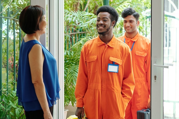 Jeunes concierges multiethniques souriants en combinaisons orange debout à l'entrée de la maison et venant dans la maison des clients, femme mûre les accueillant