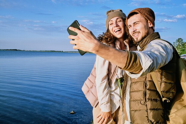 Jeunes dates joyeuses debout les uns par les autres et regardant la caméra du smartphone tout en faisant selfie sur fond de ciel bleu et d'eau