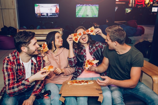 Les jeunes drôles sont assis ensemble dans la chambre et mangent de la pizza. Ils jouent avec ses pièces. Coéquipiers riant.