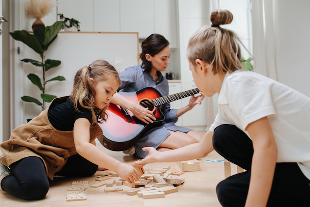 Jeunes enfants jouant sur le sol de la cuisine avec des blocs de bois pendant que maman joue de la guitare en arrière-plan