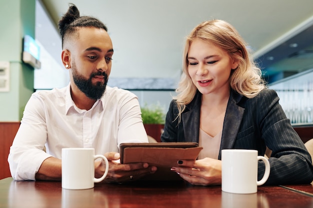 Jeunes entrepreneurs positifs buvant du café au café et lisant un article en ligne sur le projet sur lequel ils travaillaient