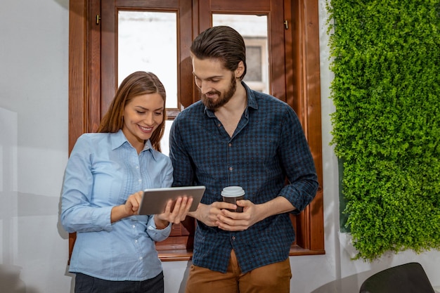 Jeunes entrepreneurs souriants prenant une pause-café et regardant quelque chose d'intéressant sur une tablette numérique devant la fenêtre du bureau.