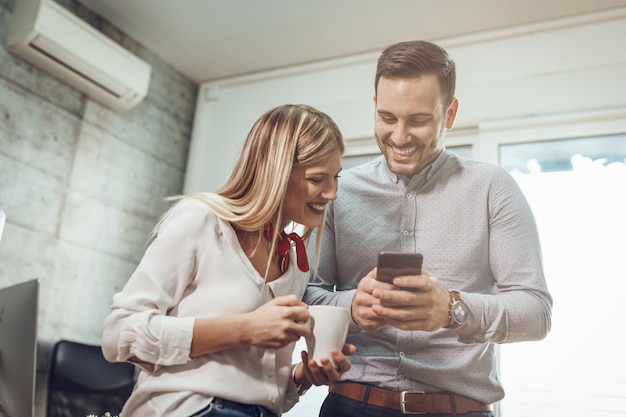 Jeunes entrepreneurs souriants prenant une pause-café et utilisant un téléphone intelligent au bureau.