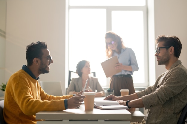 Jeunes Experts En Marketing Multiethnique Positifs Assis à Table Dans La Salle De Réunion Et Partageant Des Idées Tout En Discutant D'une Nouvelle Campagne Publicitaire