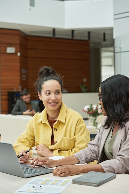 Jeunes femmes d'affaires assises à la table avec un ordinateur portable et se parlant lors d'une réunion au bureau