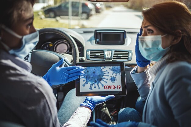 De jeunes femmes d'affaires portant des masques de protection sur le visage sont assises dans une voiture et parlent de la crise mondiale. Ils se montrent une animation d'un virus corona sur une tablette.