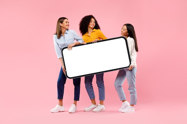 Jeunes femmes diverses tenant un smartphone géant avec une maquette pour une publicité démontrant l'espace pour votre conception