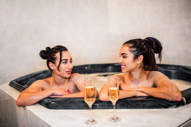 De jeunes femmes gaies s'assoient dans un bain d'hydromassage et se regardent. Ils sourient. Les mannequins ont devant eux deux verres de champaigne.