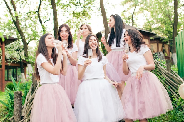 Photo jeunes femmes souriantes dans des jupes roses puder similaires et des t-shirts blancs. filles buvant du champagne dans le parc du printemps avec des arbres fruitiers en fleurs. enterrement de vie de jeune fille, célébration pré-mariage