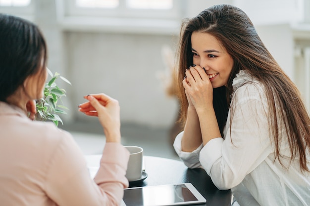 Jeunes filles insouciantes brune amis en tenue décontractée avec des tasses de thé et partage des nouvelles dans un café lumineux ou une cuisine avec des plantes vertes sur la table