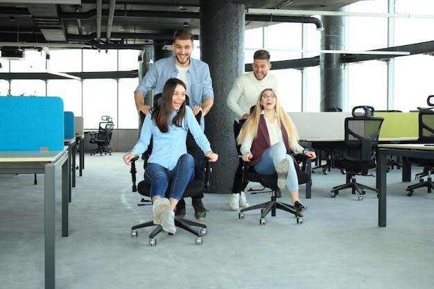 Jeunes gens d'affaires joyeux dans des vêtements décontractés intelligents s'amusant en courant sur des chaises de bureau et en souriant.