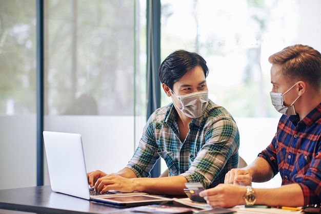 Jeunes gens positifs dans des masques médicaux assis à la table de bureau et discutant de la manière de développement des affaires en ligne