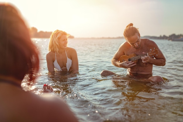 Des jeunes heureux qui passent un bon moment ensemble à la plage. Ils sont assis dans l'eau et l'homme joue du ukulélé et chante. Coucher de soleil sur l'eau.