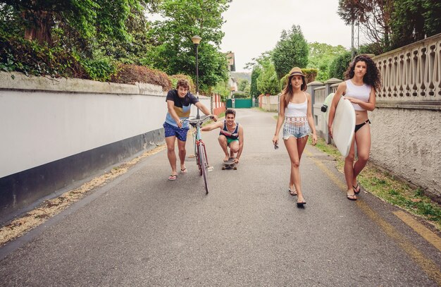 Des jeunes heureux en vêtements d'été marchant le long de la route et s'amusant avec une planche à roulettes et un vélo. Concept de mode de vie d'été.