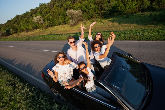 Des jeunes hommes élégants sont assis, levant les mains et souriant dans un cabriolet noir sur la route de campagne par une journée ensoleillée. .