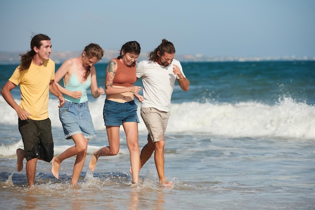 Jeunes hommes et femmes gais courant sur la plage et éclaboussant de l'eau par une journée ensoleillée