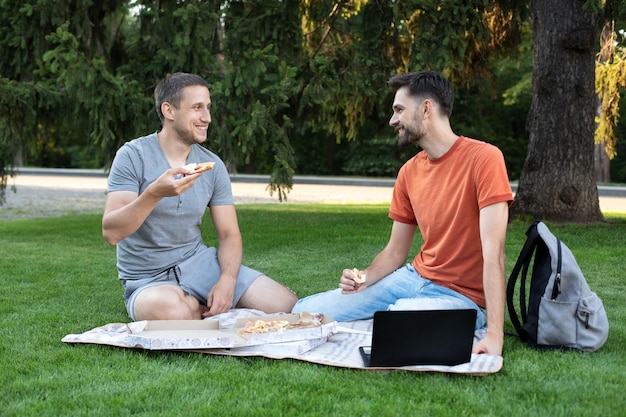 Les jeunes hommes mangent de délicieuses pizzas, parlent et rient des blagues. Des amis assis sur la nature à l'extérieur mangent de la pizza en pique-nique.