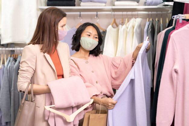 Jeunes jolies femmes interculturelles portant des masques et des vêtements décontractés intelligents debout près d'un rack avec un nouvel assortiment de vêtements chauds et choisissant des robes