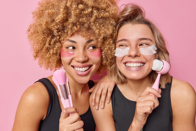 De jeunes jolies femmes positives appliquent des patchs de beauté sous les yeux pour le traitement de la peau utilisent des pinceaux cosmétiques et un masseur de visage prennent soin du visage portent un t-shirt noir décontracté isolé sur fond rose